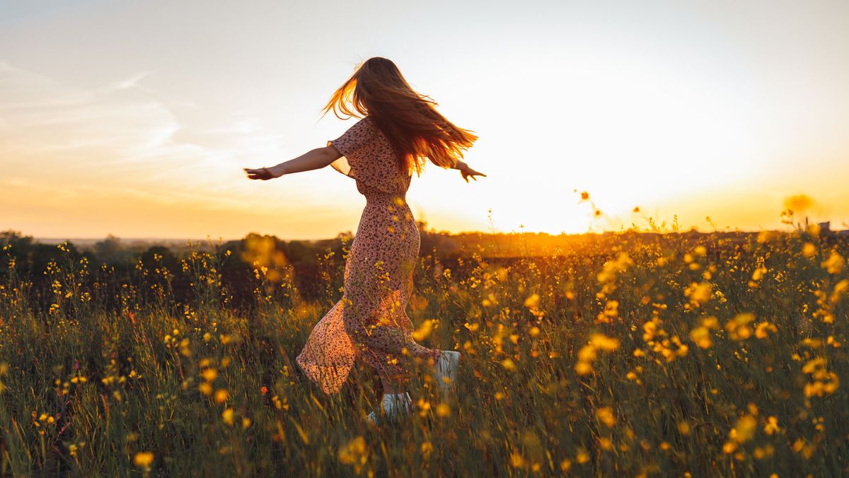 Young woman in long dress dances with joy in a rapeseed yellow field at sunset
