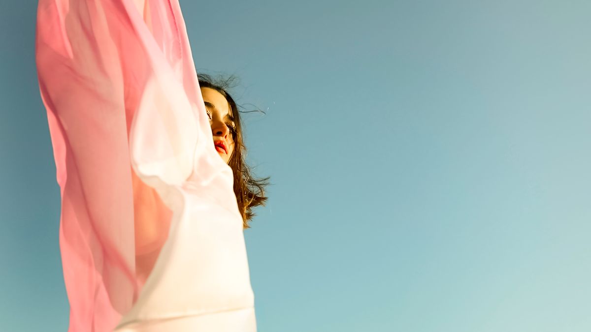 The face of a young woman peeking out from behind a pink veil that flows in the wind, against the blue sky